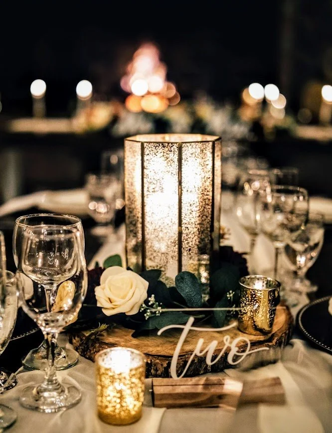 Candlelit wedding tablescape with glass lantern centerpiece, white rose, and “two” table number among wine glasses.