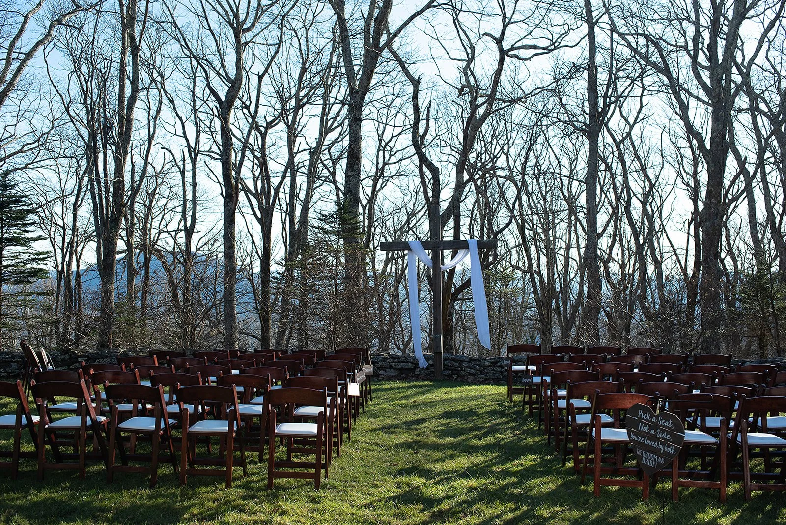 Outdoor ceremony site with rows of chairs facing a wooden arbor draped with white fabric.