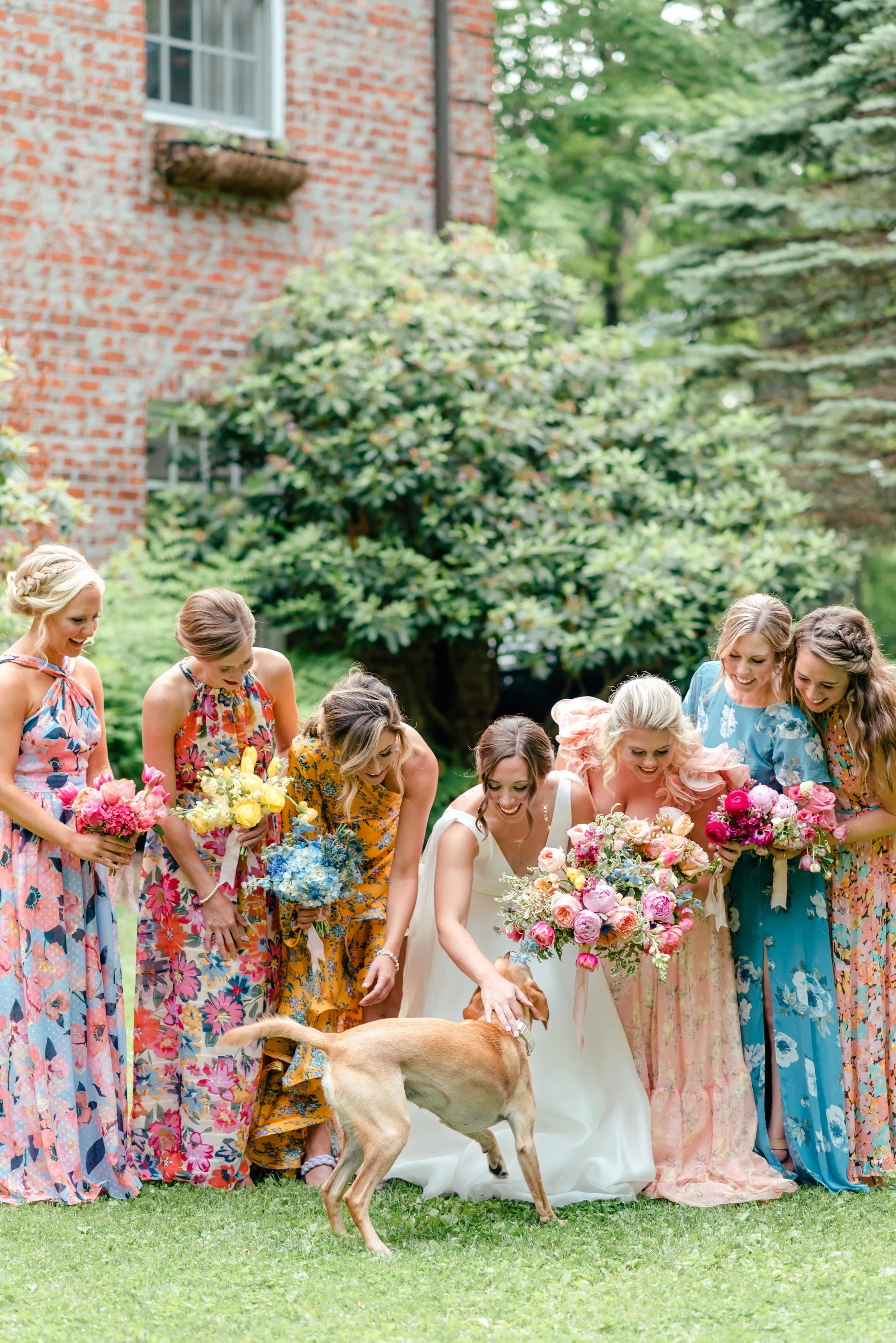 Bride and bridesmaids in colorful floral dresses smiling at a dog during outdoor wedding portraits.