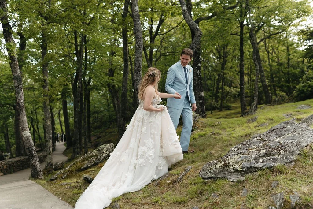 Bride and groom walking together through a wooded mountain trail with rocks and greenery around them.