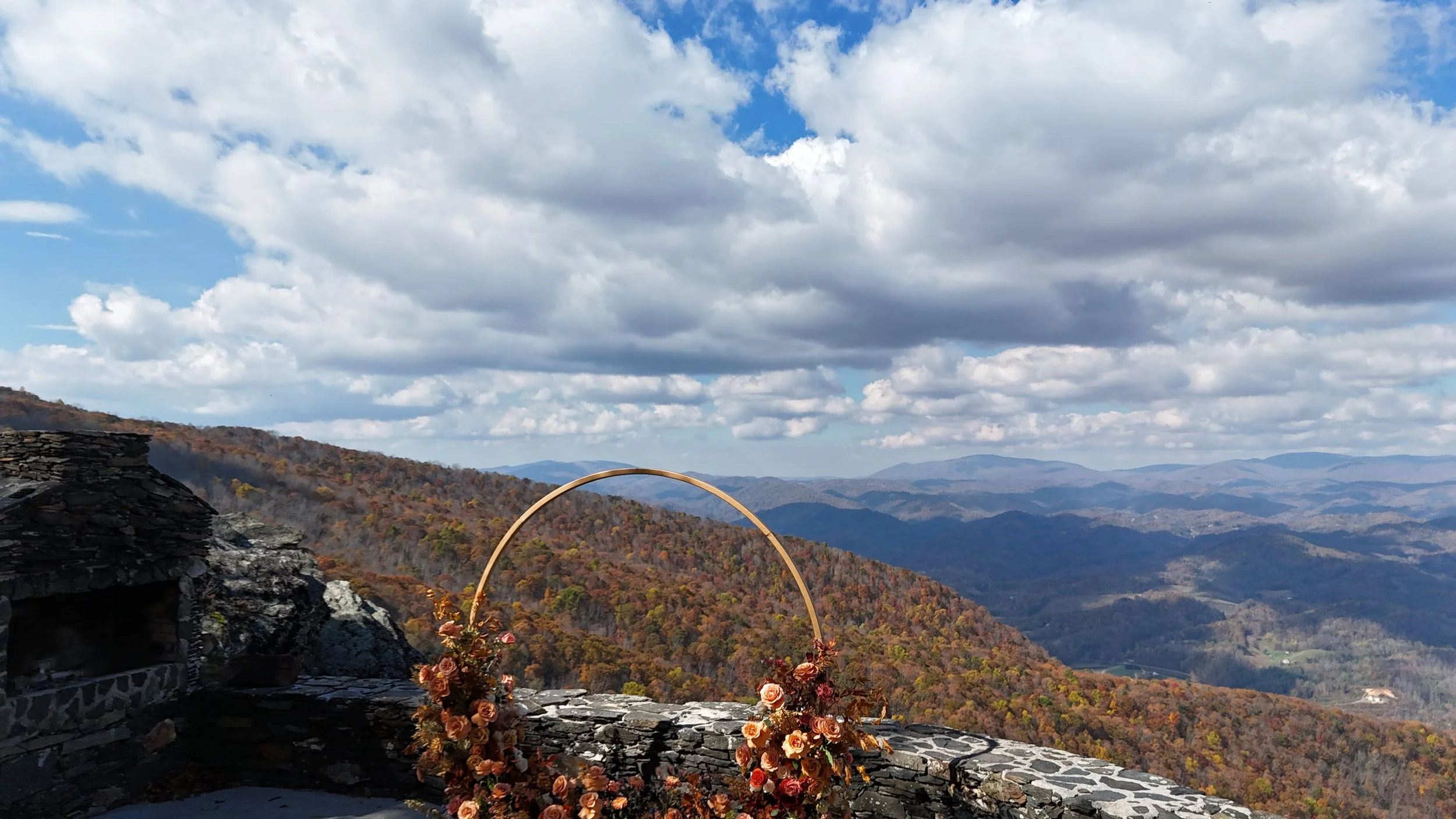 Distant aerial view of a small ceremony terrace perched on a rocky cliff, surrounded by colorful autumn forest.