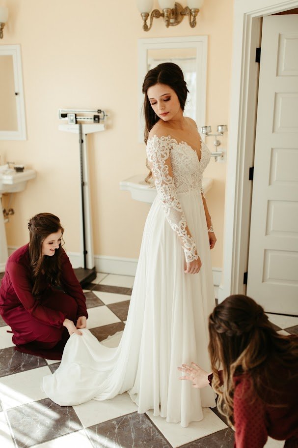Bride getting ready in a long sleeve lace wedding dress while attendants adjust the gown.