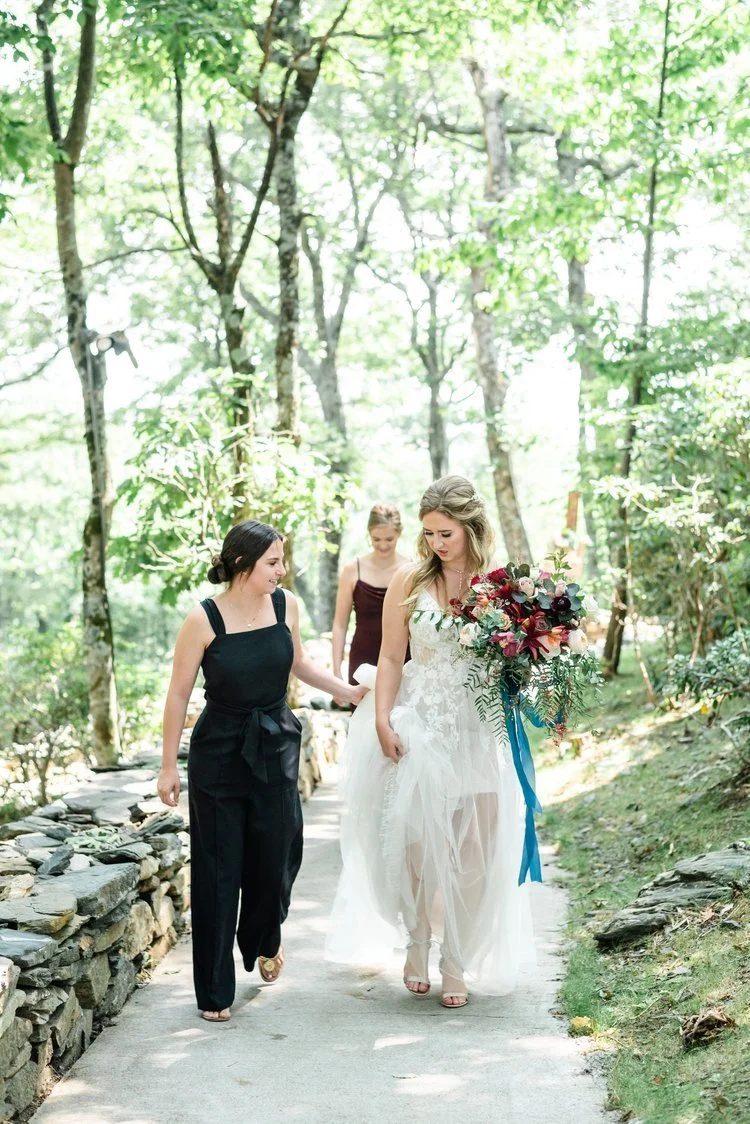 A smiling bride in a white lace dress walks along a stone path outdoors, holding a colorful bouquet, accompanied by two attendants.