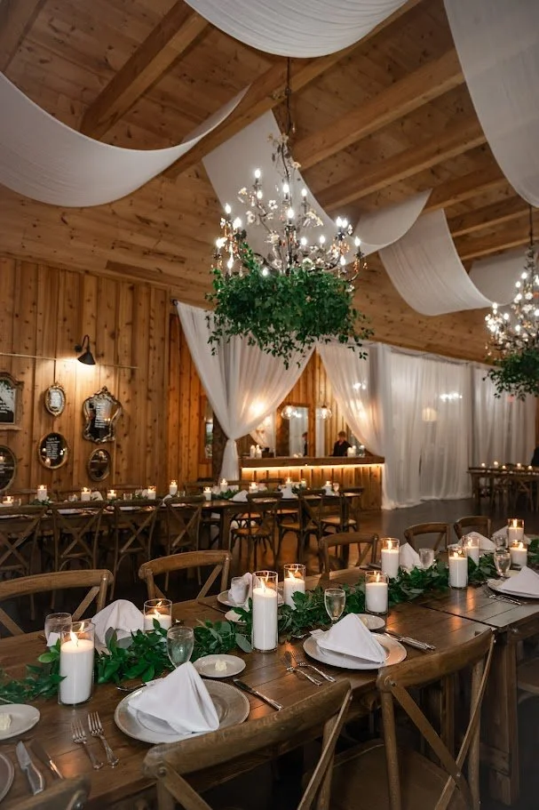 Rustic barn wedding reception with long wooden tables, candlelight, chandeliers, and a draped ceiling