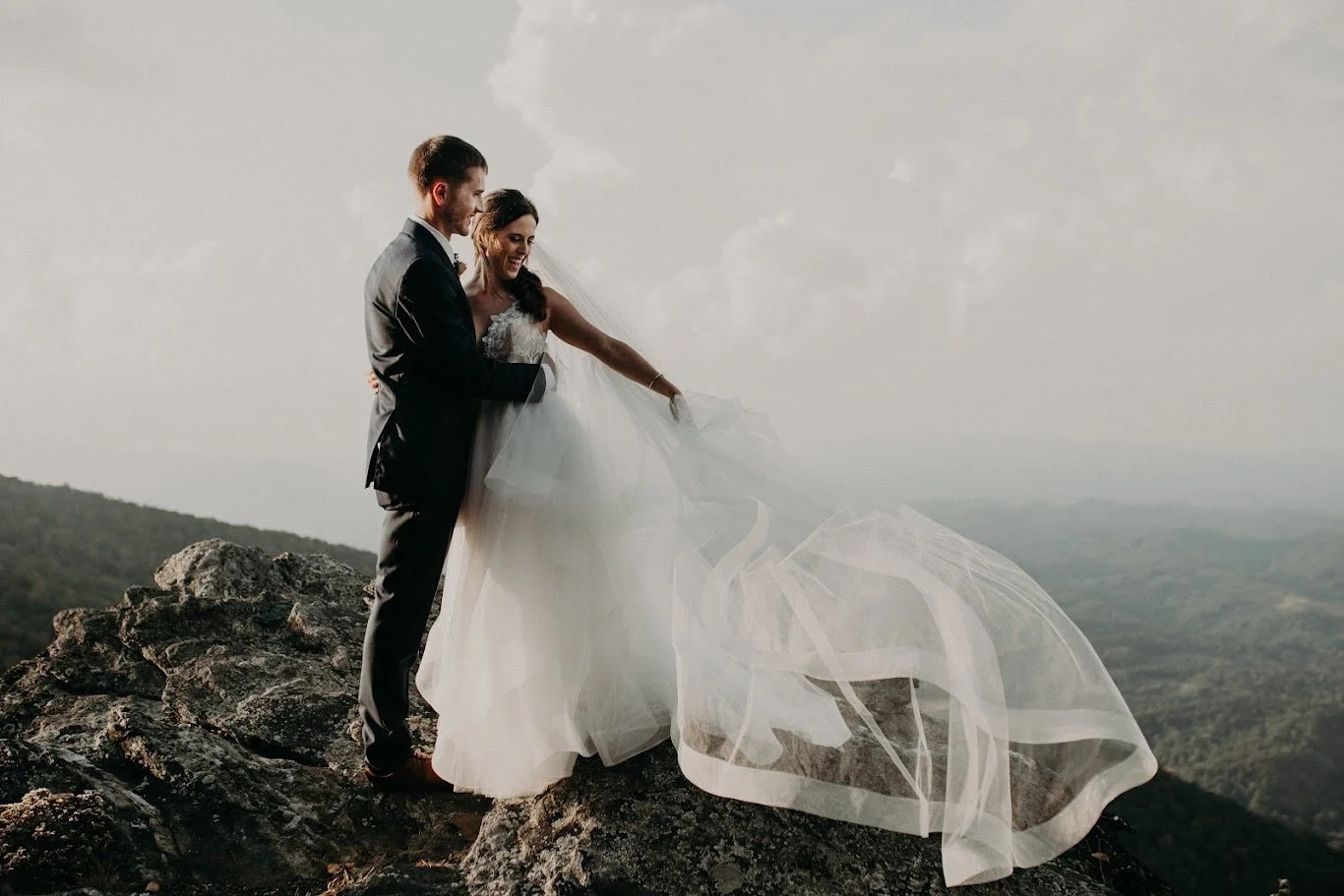 Bride and groom standing together on a mountain cliff with the bride’s veil flowing in the wind.