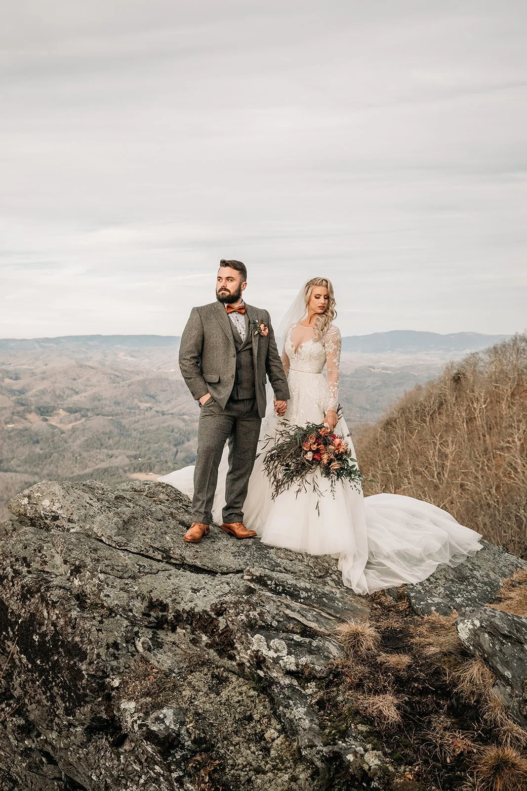 Bride and groom standing on a rocky mountain overlook with distant blue ridge views behind them.