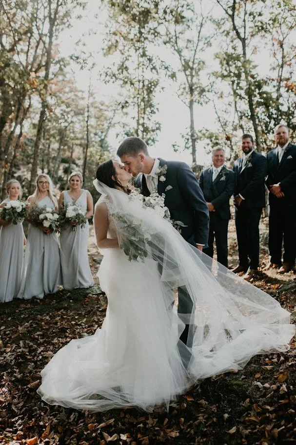 Bride and groom sharing a dip kiss during an outdoor woodland wedding ceremony with their wedding party nearby.