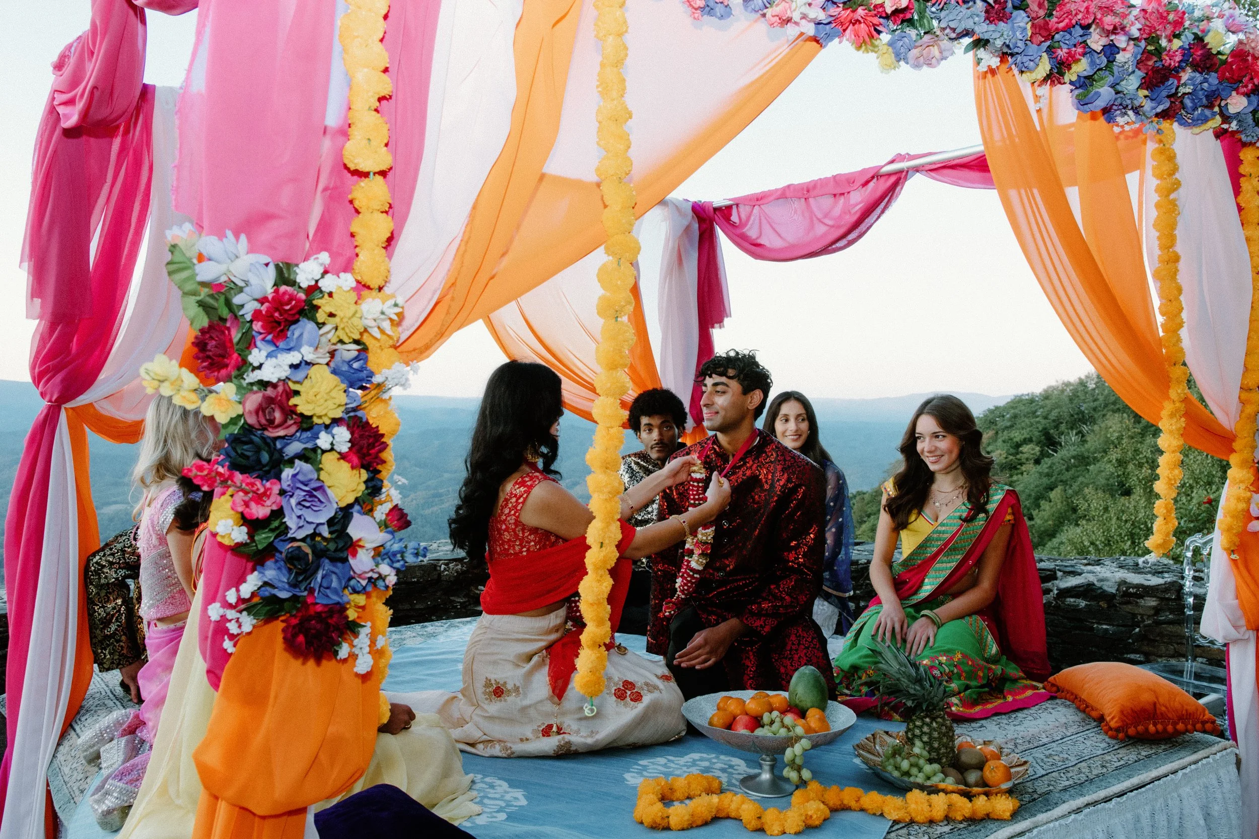 Couple sits under a bright draped canopy while a woman places a garland; guests and fruit offerings surround them.