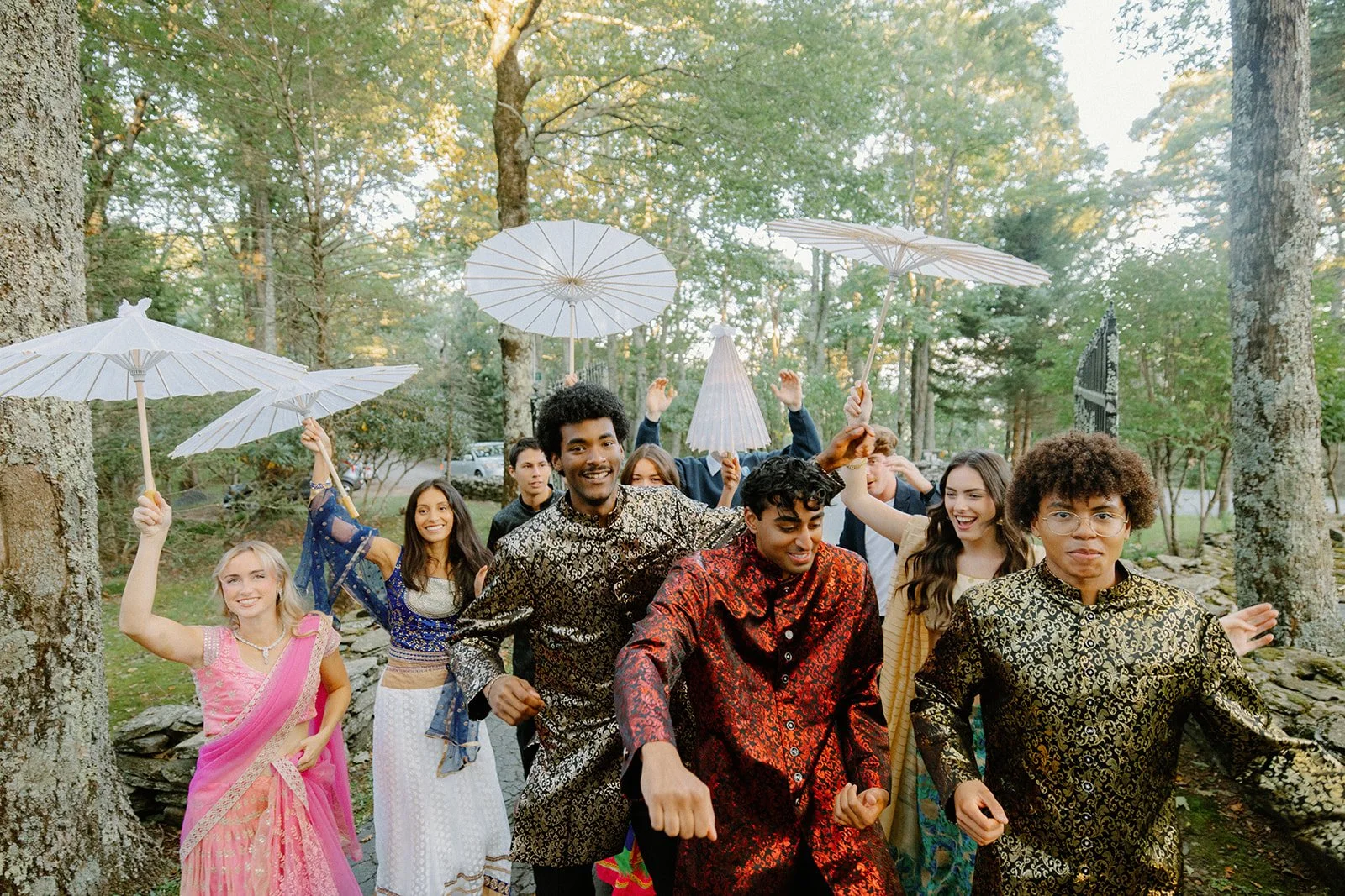 Group dancing outdoors in a forest during an Indian wedding celebration, holding white parasols