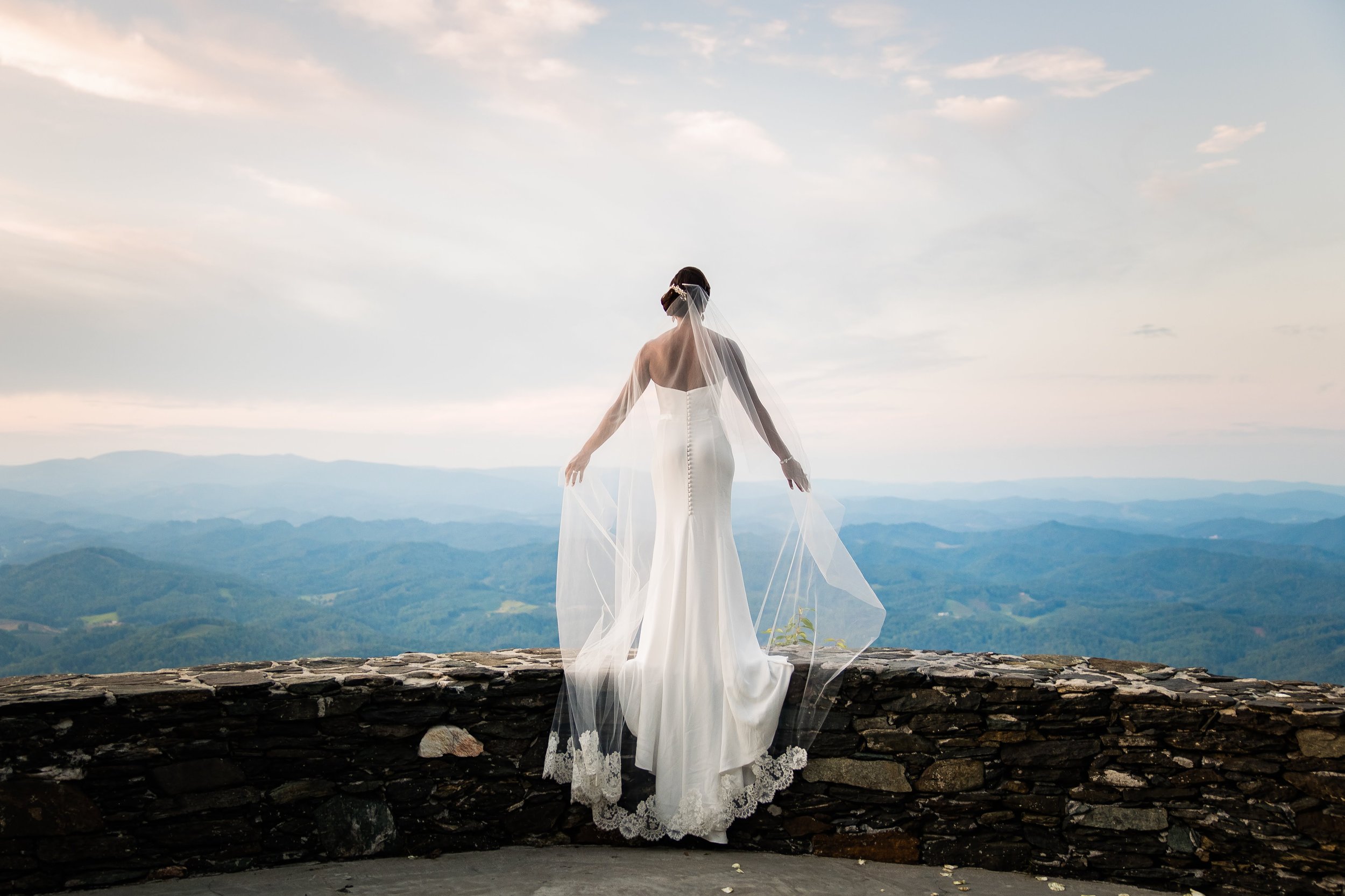 Bride in a lace-trim veil and fitted wedding gown standing on a stone wall, overlooking the Blue Ridge Mountains