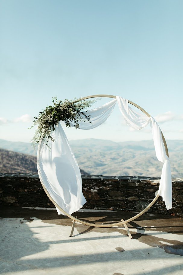 Circular mountain wedding ceremony arch with white draping and greenery overlooking distant hills.