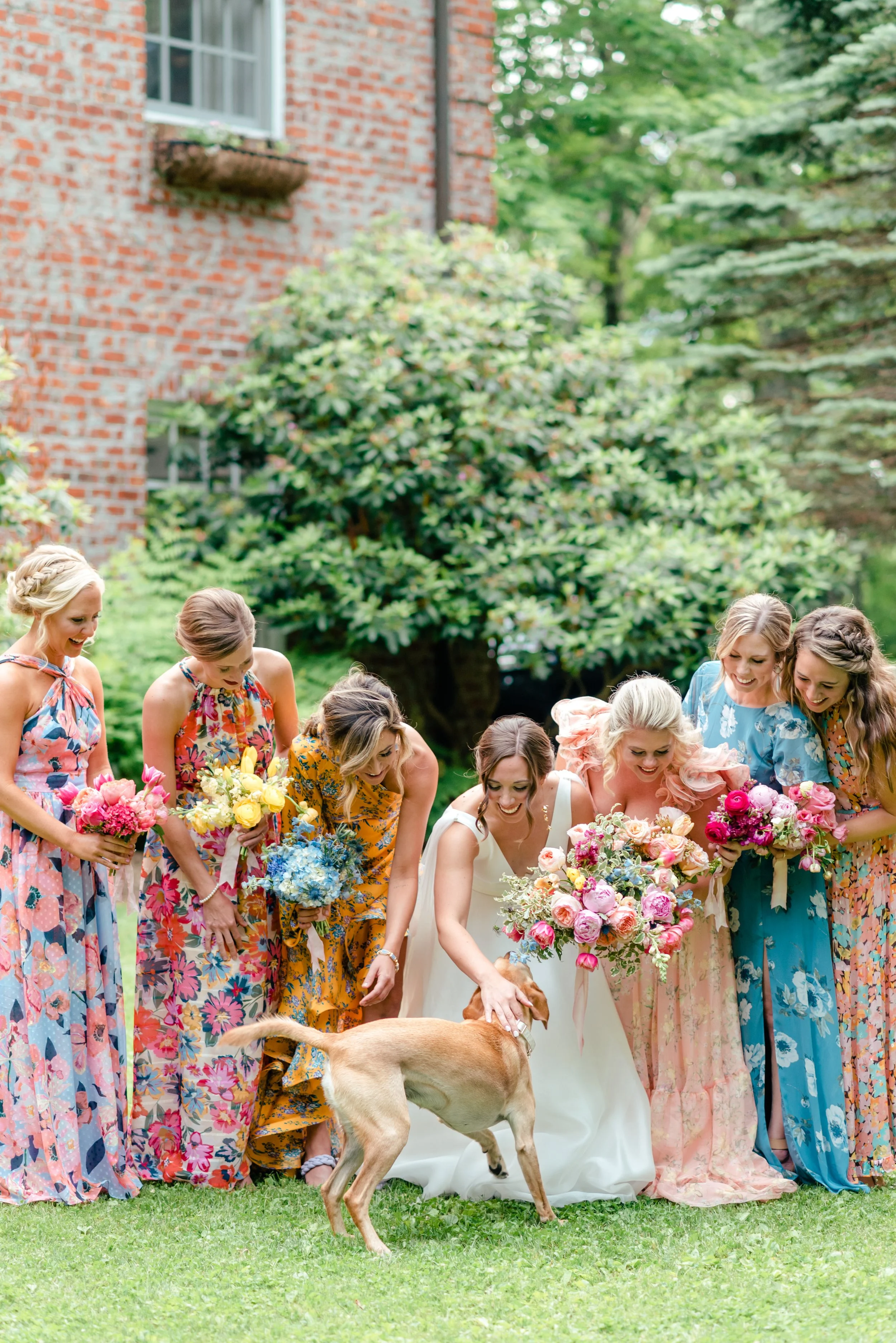 Bride and bridesmaids in colorful floral dresses smiling and petting a dog on the lawn during an outdoor wedding celebration.