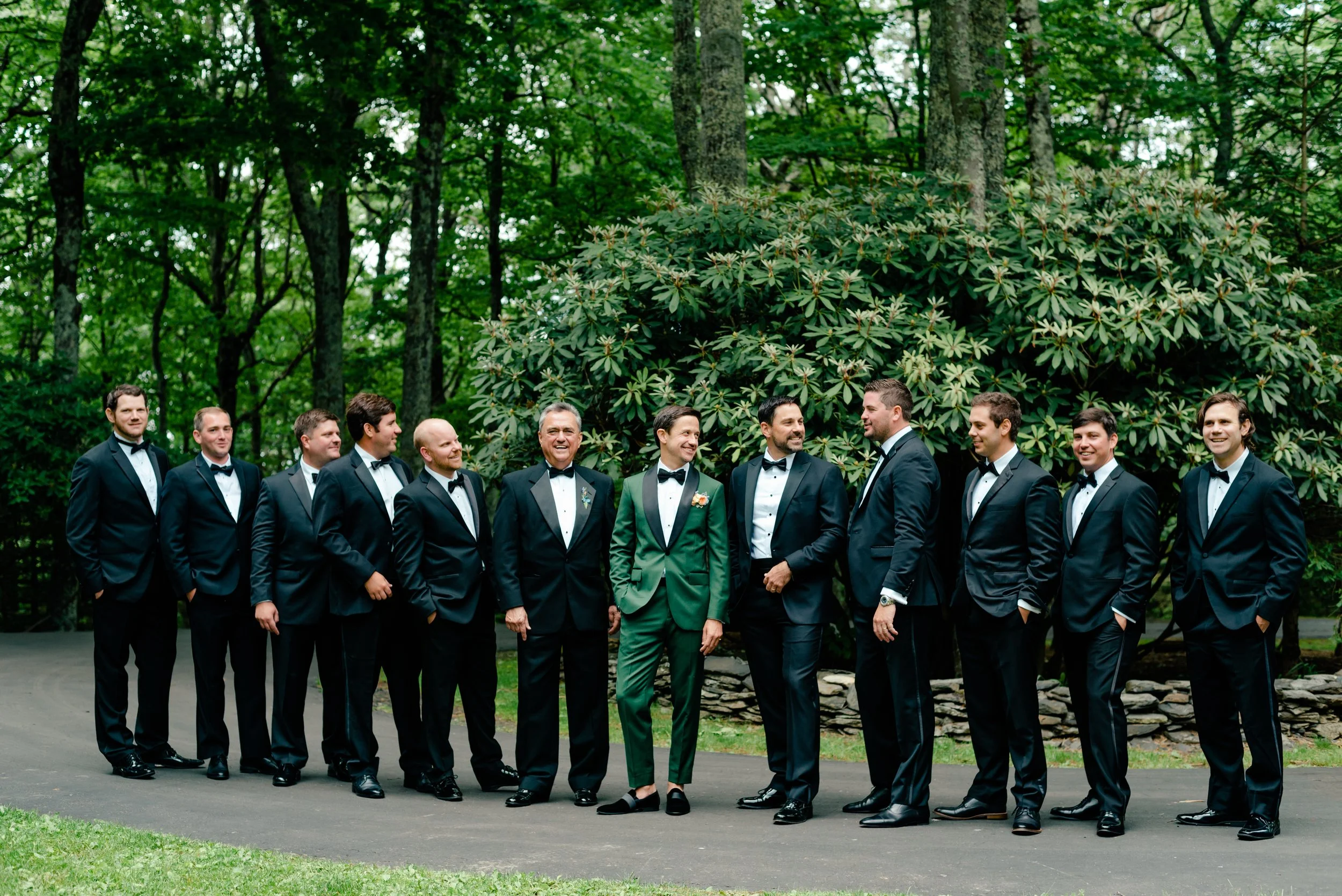 Groom in a green suit standing with groomsmen in black tuxedos for an outdoor wedding portrait.