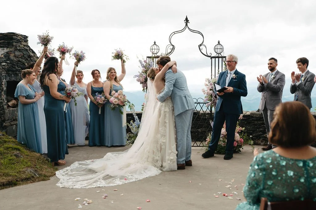 Bride and groom kissing during their outdoor wedding ceremony beneath a pastel floral arch at a mountain venue.