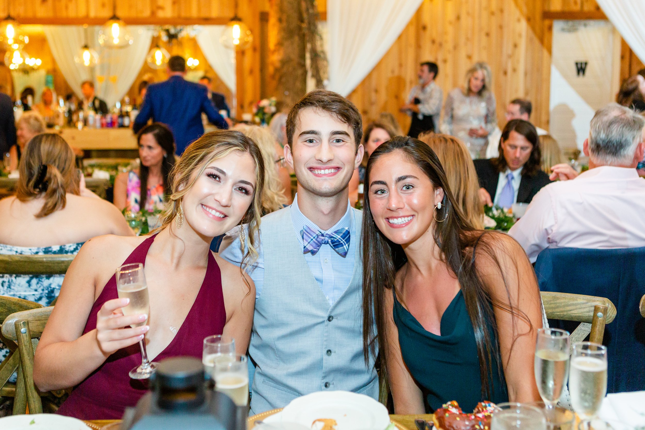 Three wedding guests smiling for a photo at a rustic indoor reception dinner with champagne glasses and wood-paneled walls.