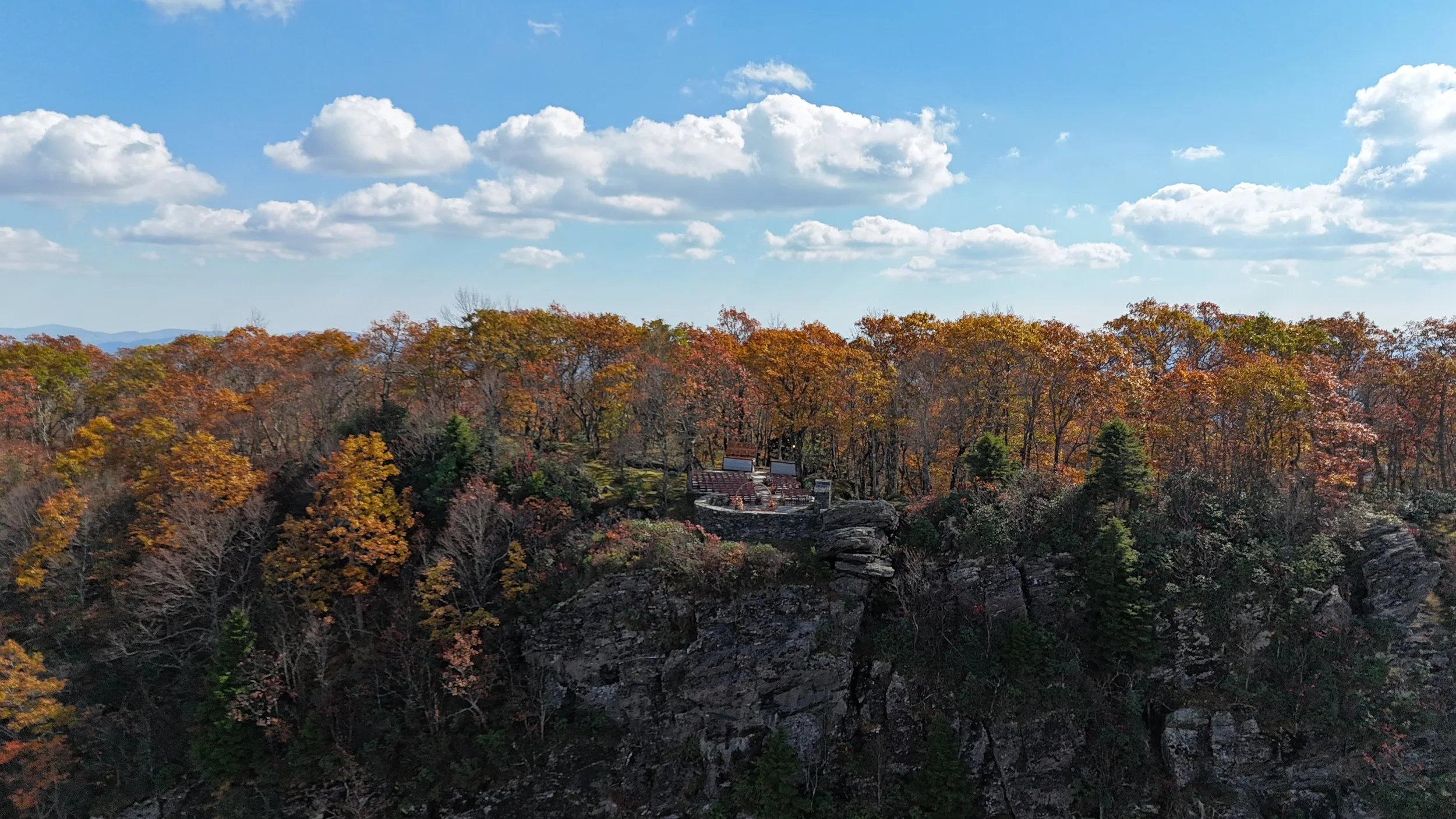 Aerial view of an outdoor ceremony site with rows of wooden chairs on a curved stone terrace among fall trees.