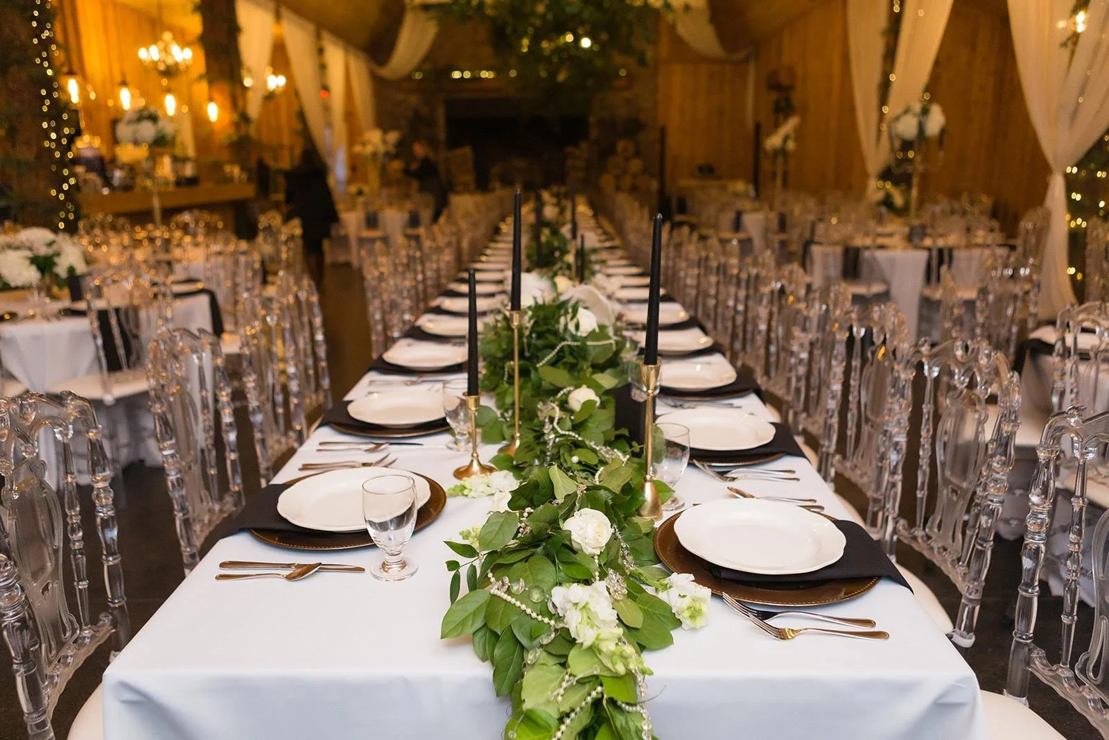 Long head table with place settings, clear chairs, and a greenery runner in a reception space.