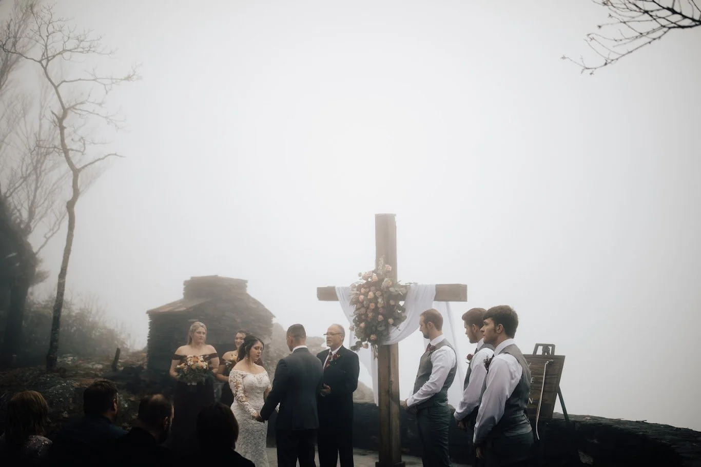 Outdoor ceremony in fog with a wooden cross at the altar and guests seated nearby.