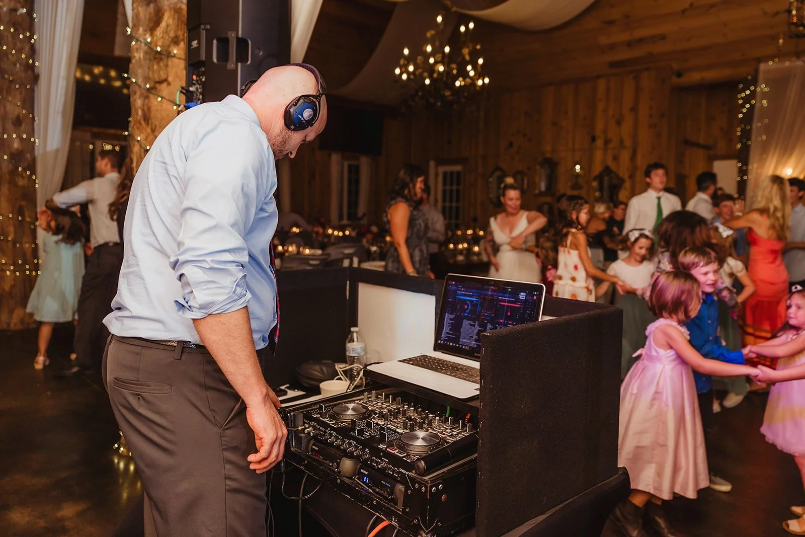 DJ wearing headphones at a booth with a laptop and mixer while wedding guests, including children, dance in the background.