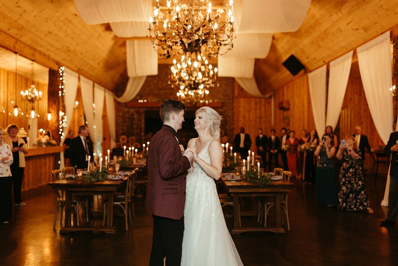Bride and groom sharing their first dance in a candlelit barn reception space with guests watching