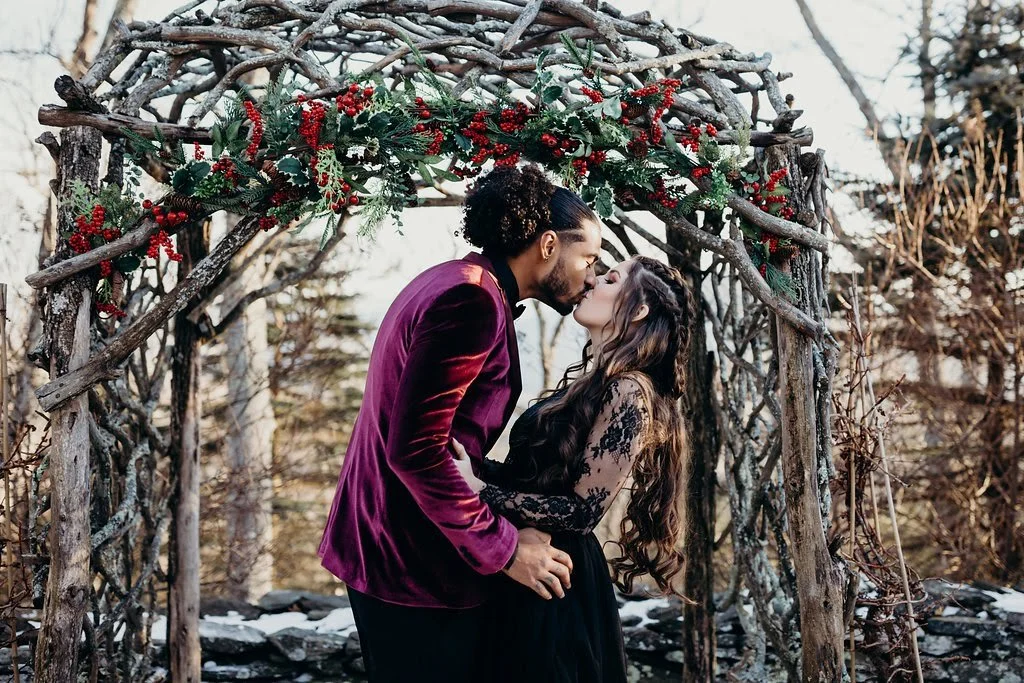 Couple kissing under a rustic wooden arch decorated with winter greenery and red berries