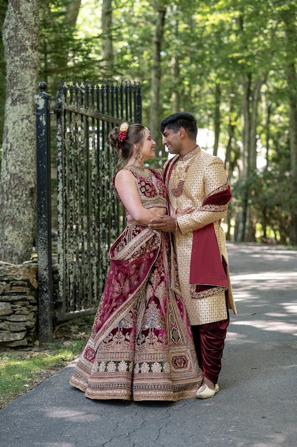 Couple in traditional South Asian outfits embrace beside an iron fence, smiling at each other in a wooded setting.
