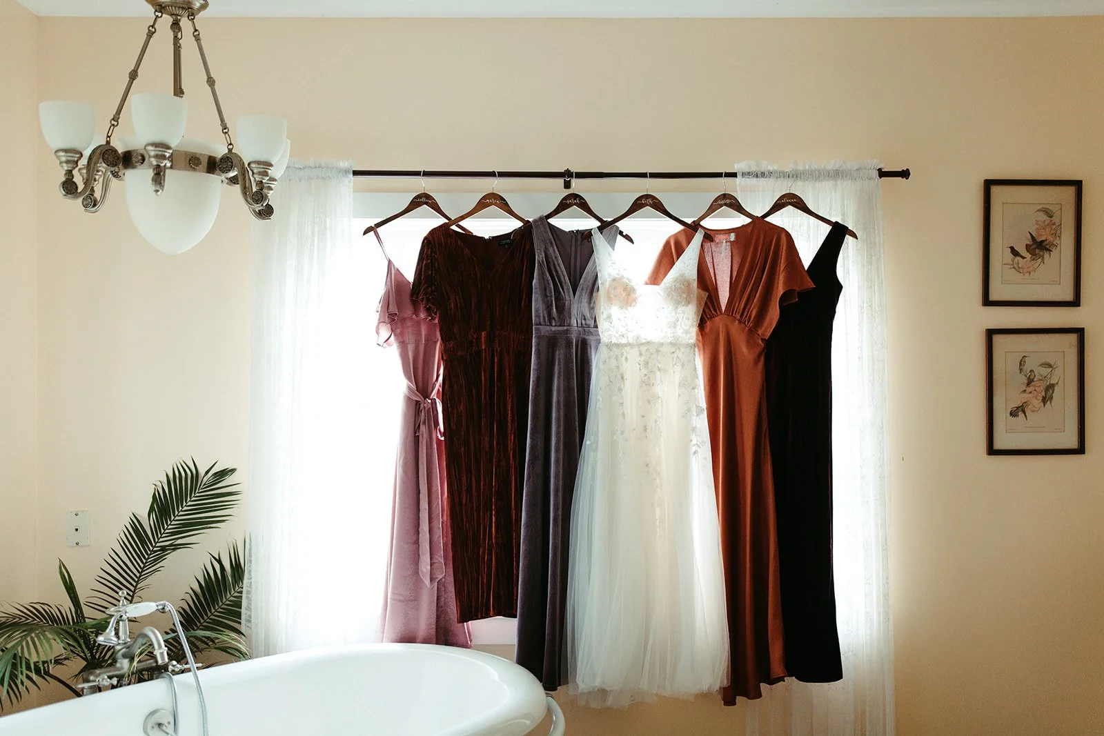Bridesmaid dresses in warm tones and a white wedding dress hanging by a bright window before the ceremony.