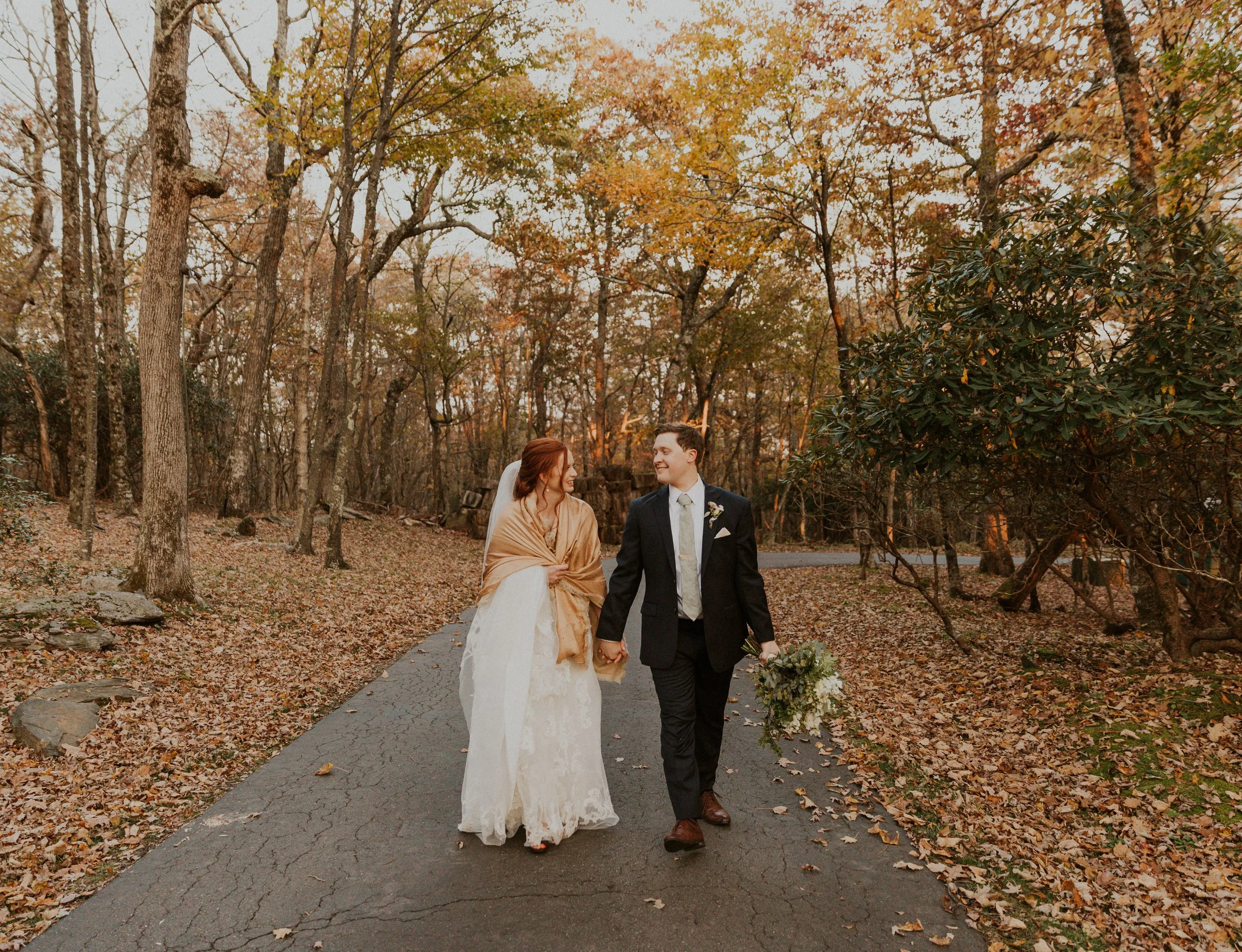 Bride and groom holding hands and walking down a quiet road surrounded by fall trees
