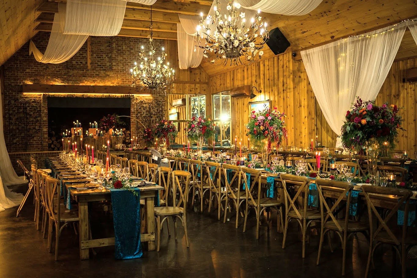 Rustic barn wedding reception with long banquet tables, chandeliers, candlelight, and white ceiling draping