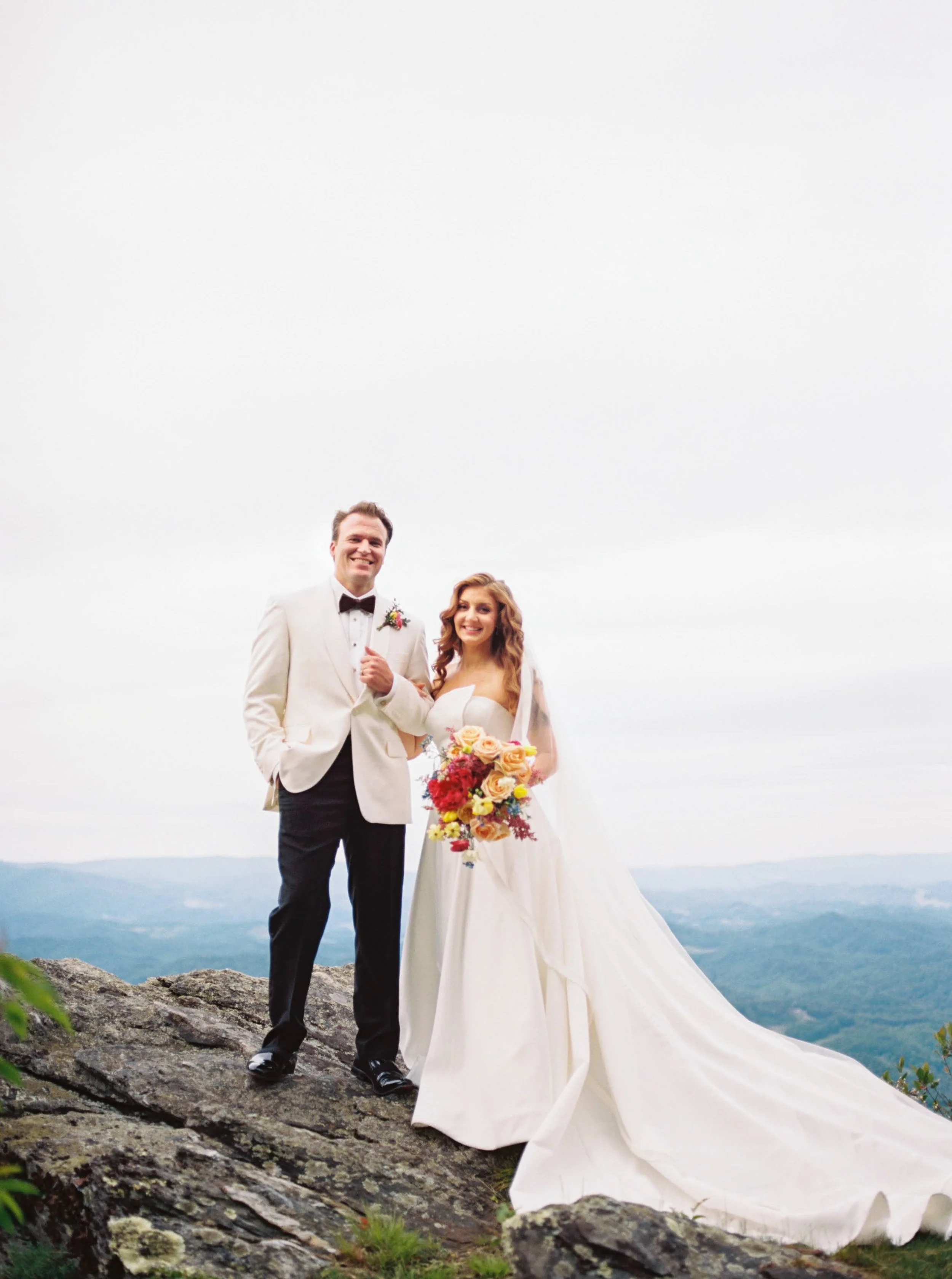 Bride and groom stand on a rocky overlook with mountain views, the groom in a white tux jacket and the bride’s long train flowing.