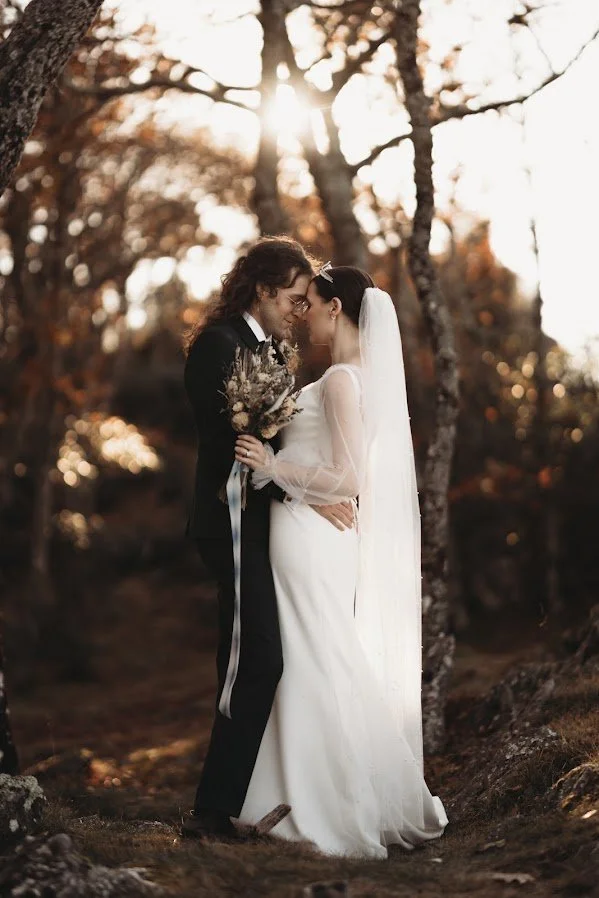 A couple in wedding attire embraces in a forest setting. The bride wears a long veil.