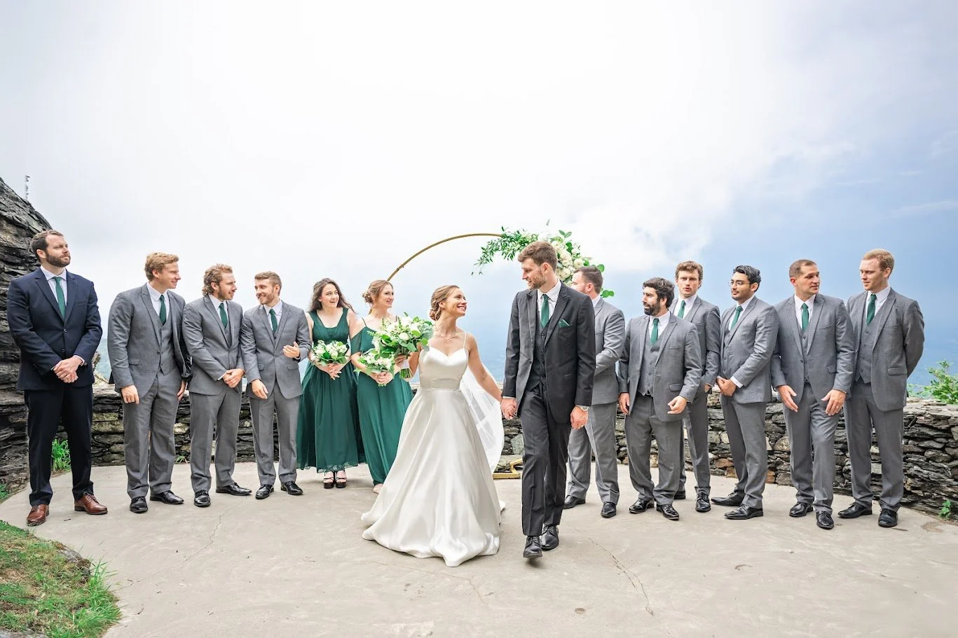 Bride and groom with wedding party in gray suits and green dresses posing by a circular arch with mountain views