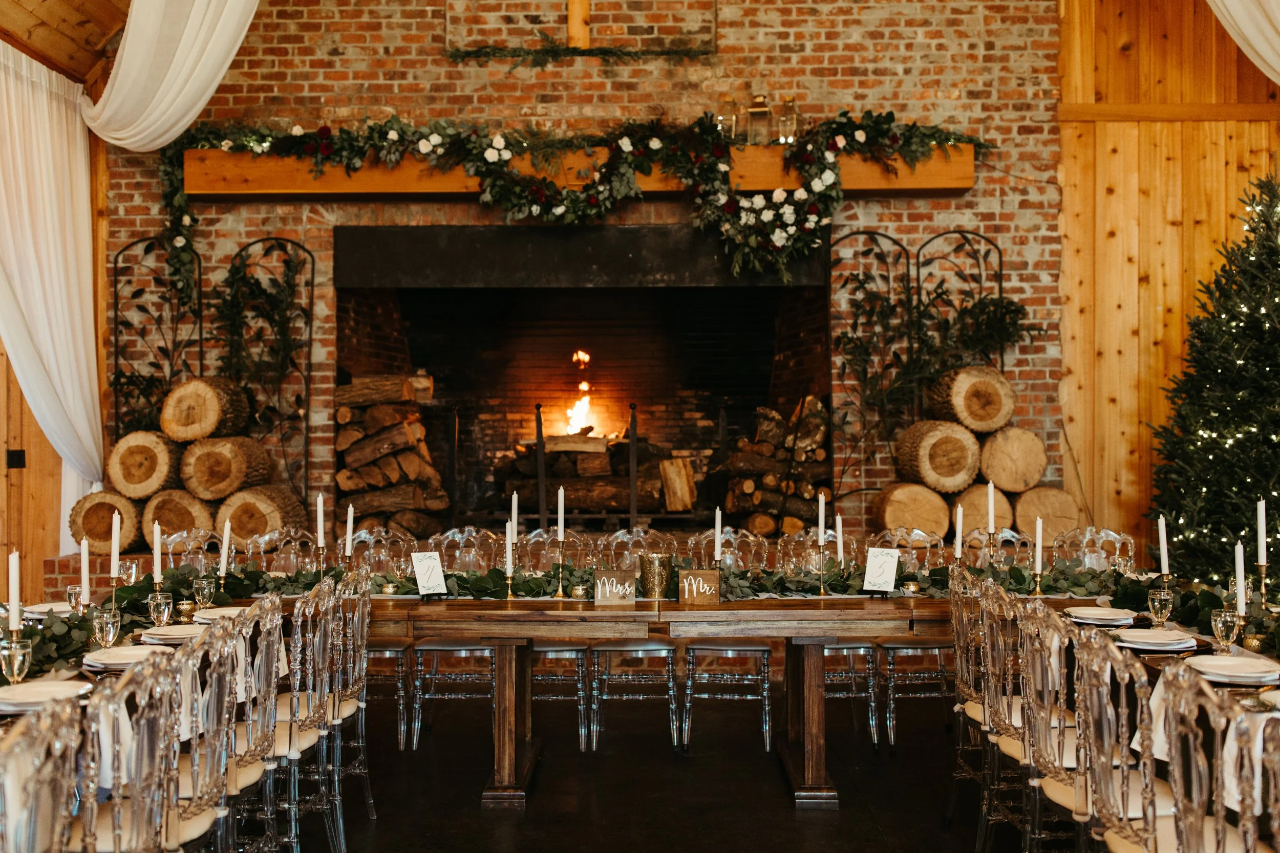 Long reception table set in front of a large brick fireplace decorated with evergreen garland and candles