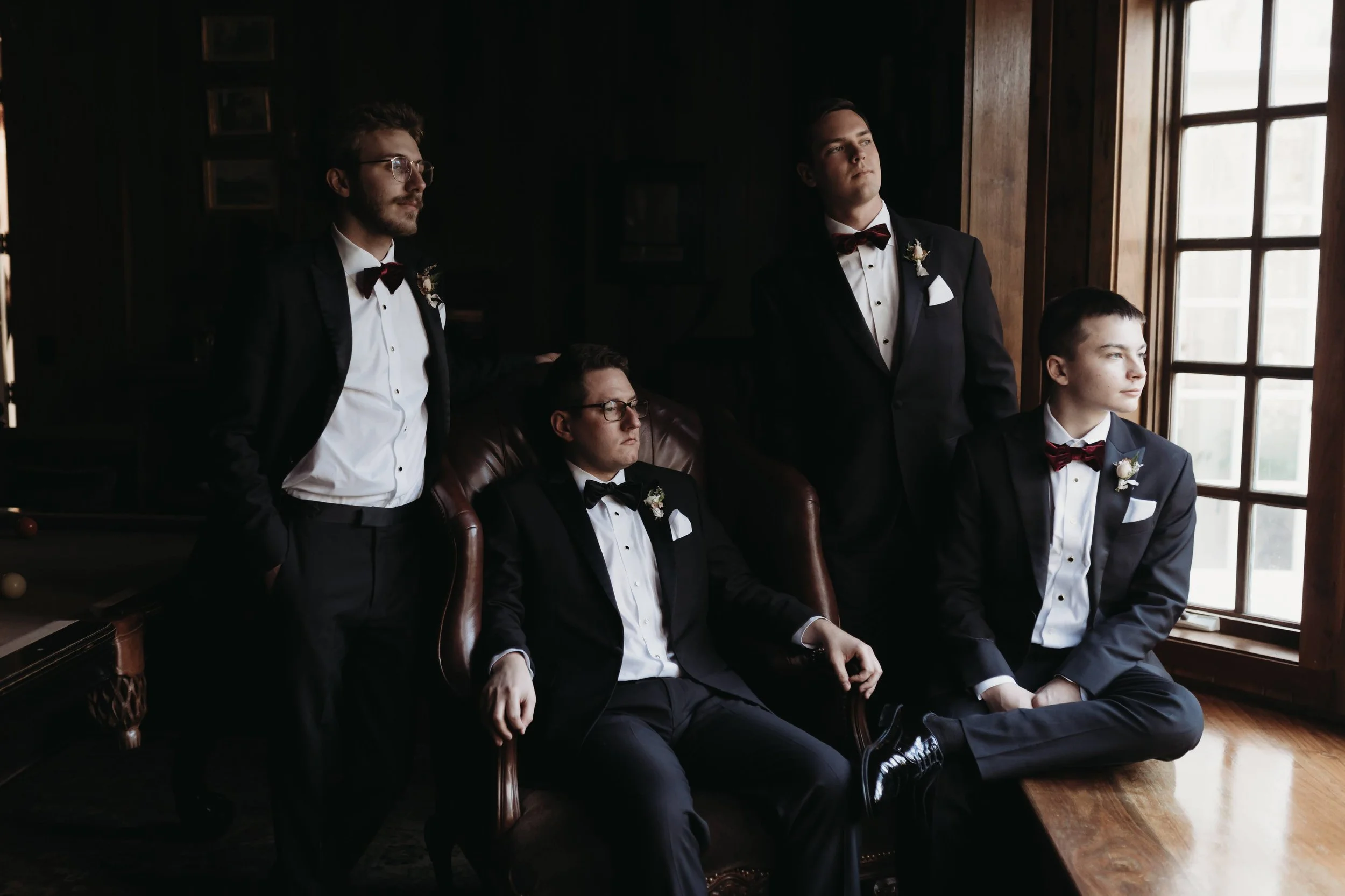 Groomsmen in tuxedos posing by a window in moody indoor light while getting ready.
