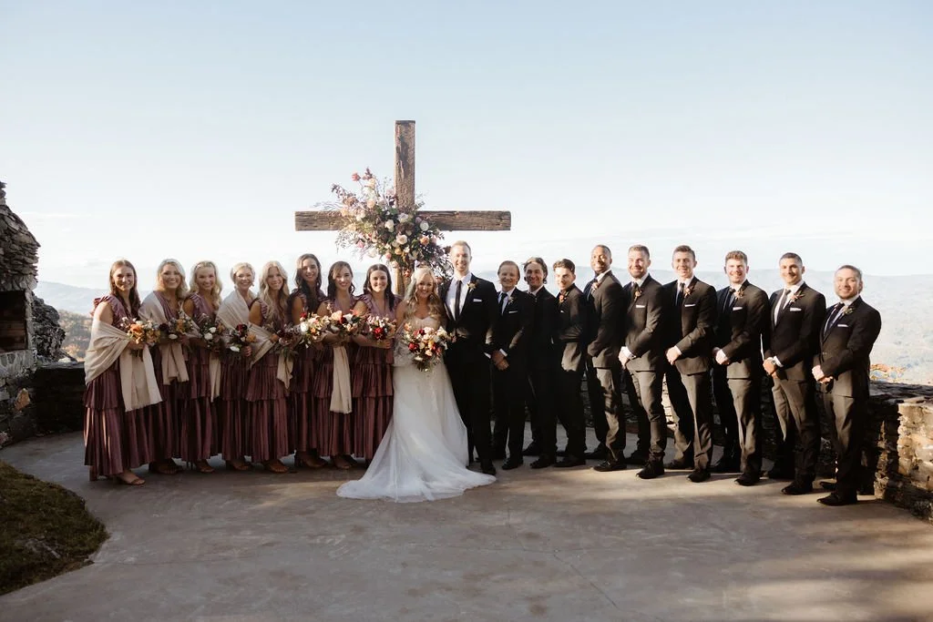 Large wedding party poses outdoors in front of a wooden cross with floral decor at a mountain overlook.