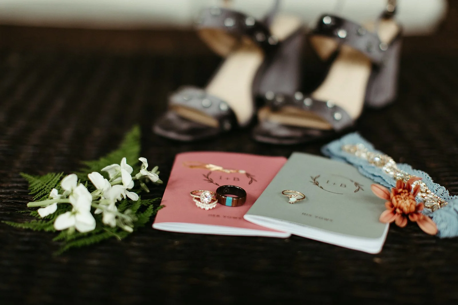 Wedding detail flat lay with vow books, rings, bridal shoes, flowers, and accessories arranged on a dark surface.