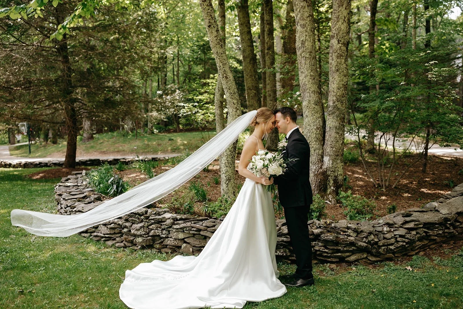 Bride and groom posing for a romantic veil portrait beside a stone wall in a wooded garden setting.