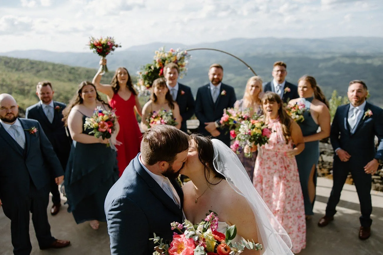 Newlyweds sharing a kiss with their wedding party standing behind them against a mountain backdrop.