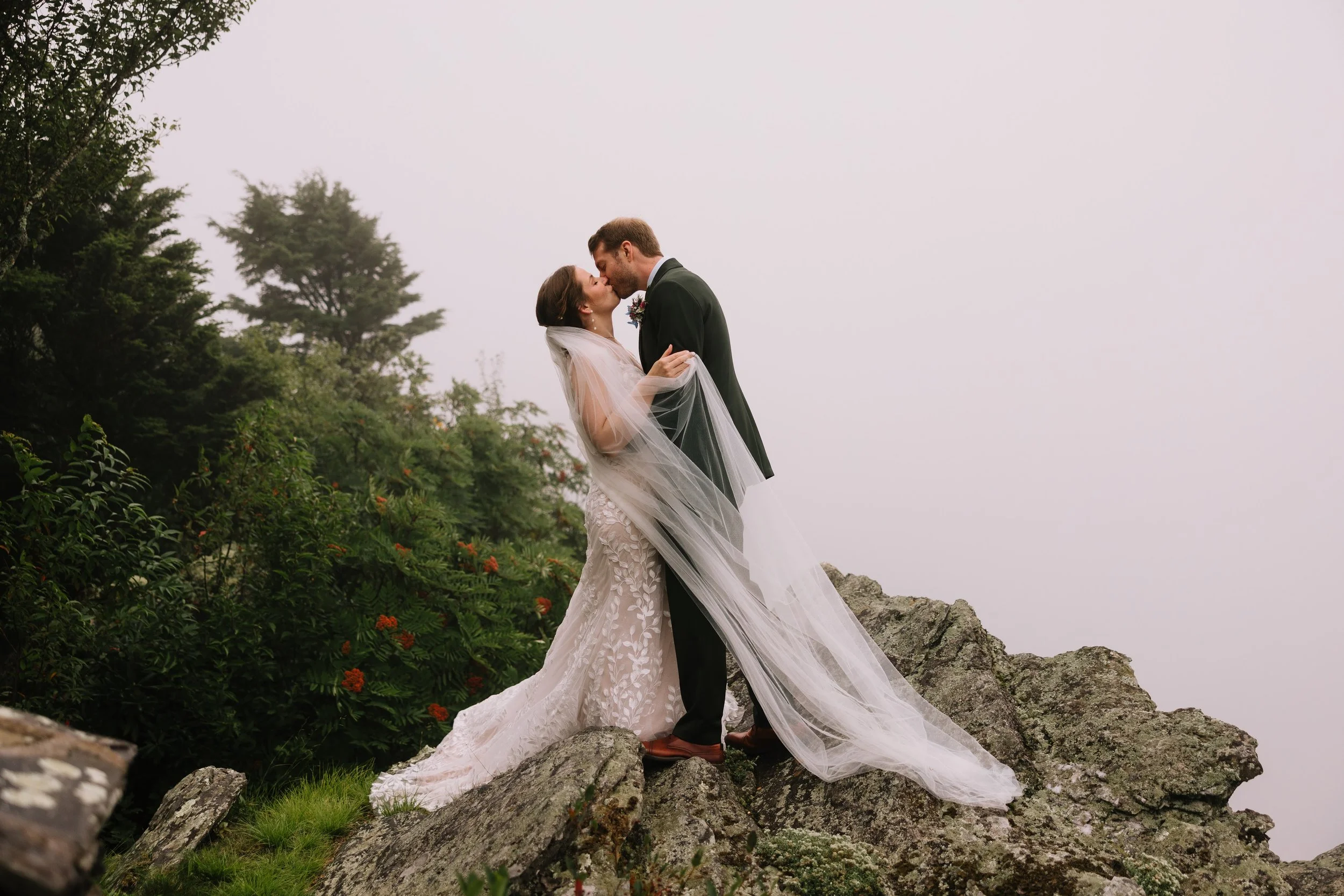 Bride and groom kissing on rocky terrain with a long veil during a foggy mountain wedding portrait.