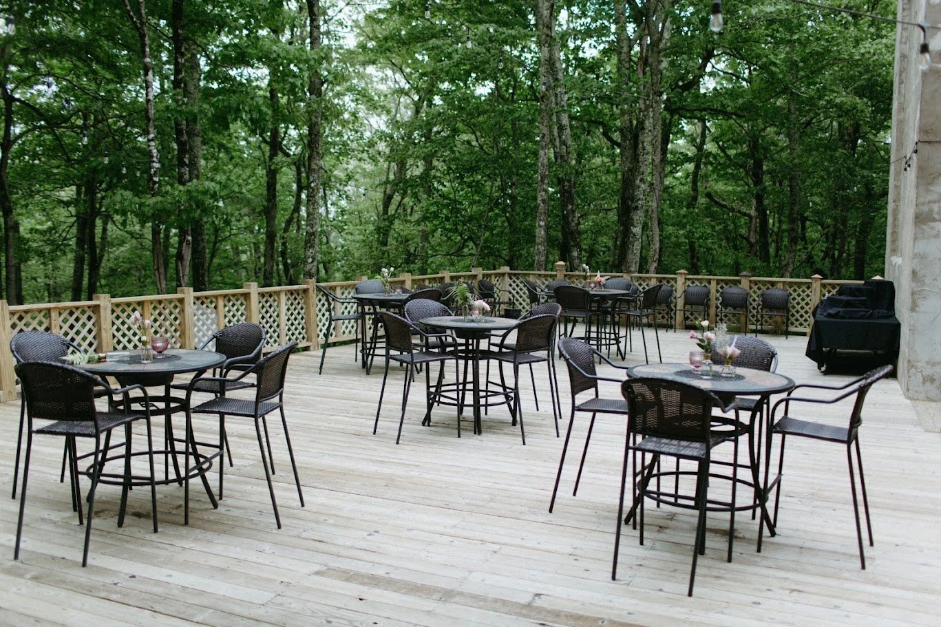Outdoor wedding reception area with multiple round tables and black chairs set up on a wooden deck surrounded by trees.
