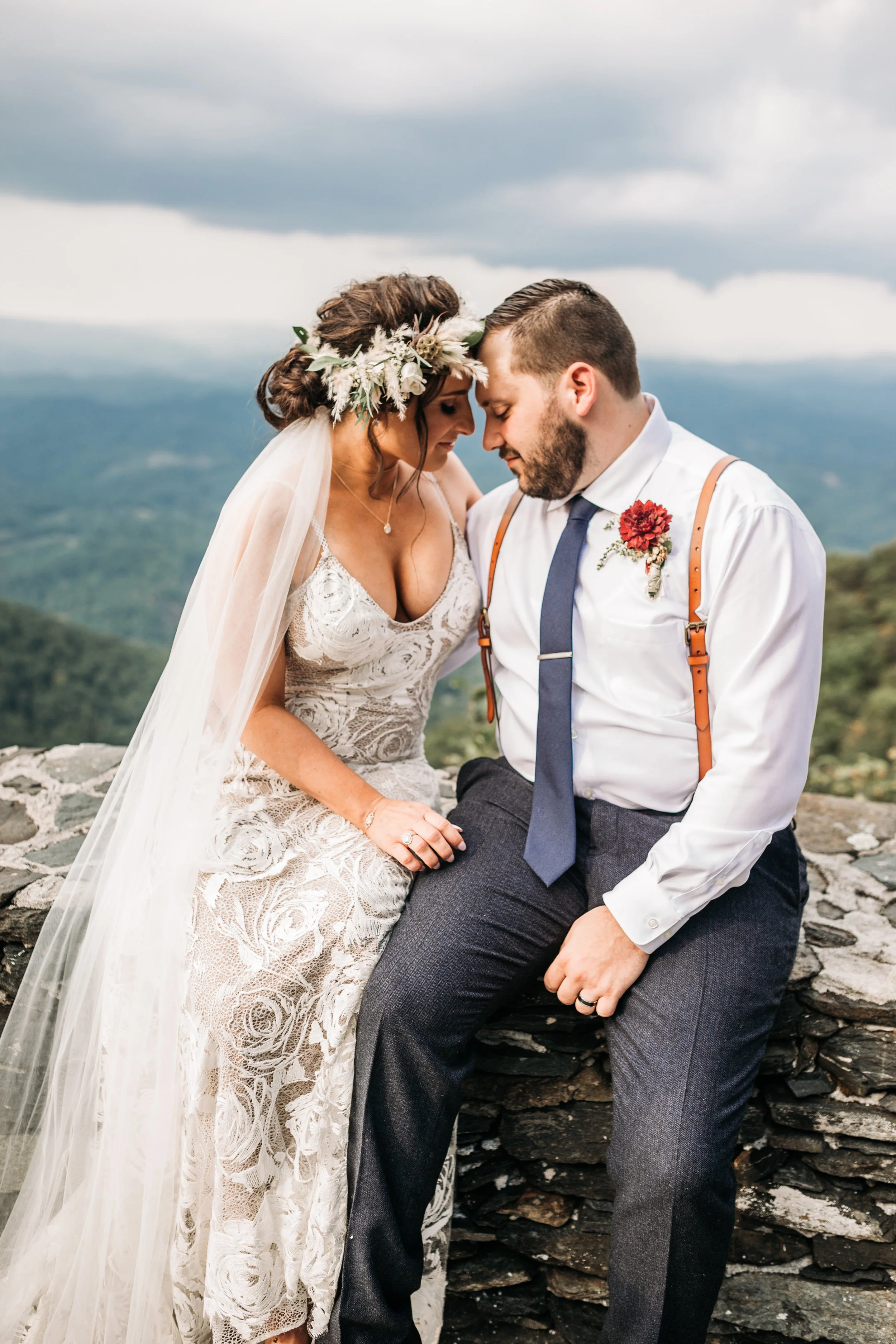 Bride and groom sitting together on a stone wall with mountains behind them, bride wearing a veil and floral crown.