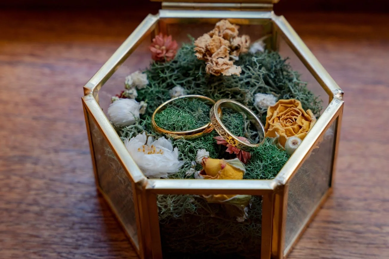 Wedding rings displayed in a hexagon glass terrarium ring box with moss and small succulents on a wooden surface.