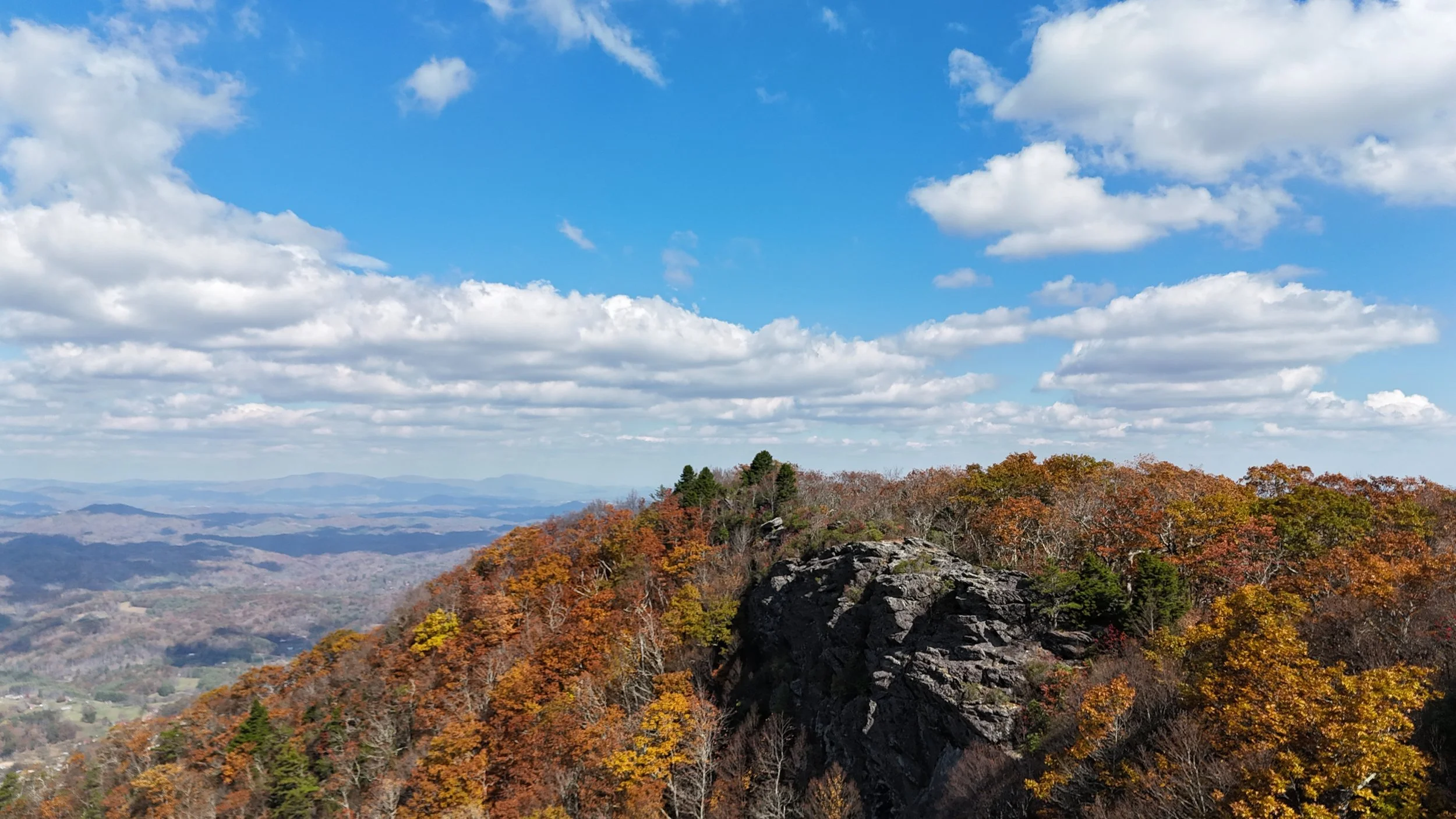 Rocky ridge lined with autumn trees under a bright blue sky with scattered clouds, overlooking distant valleys.