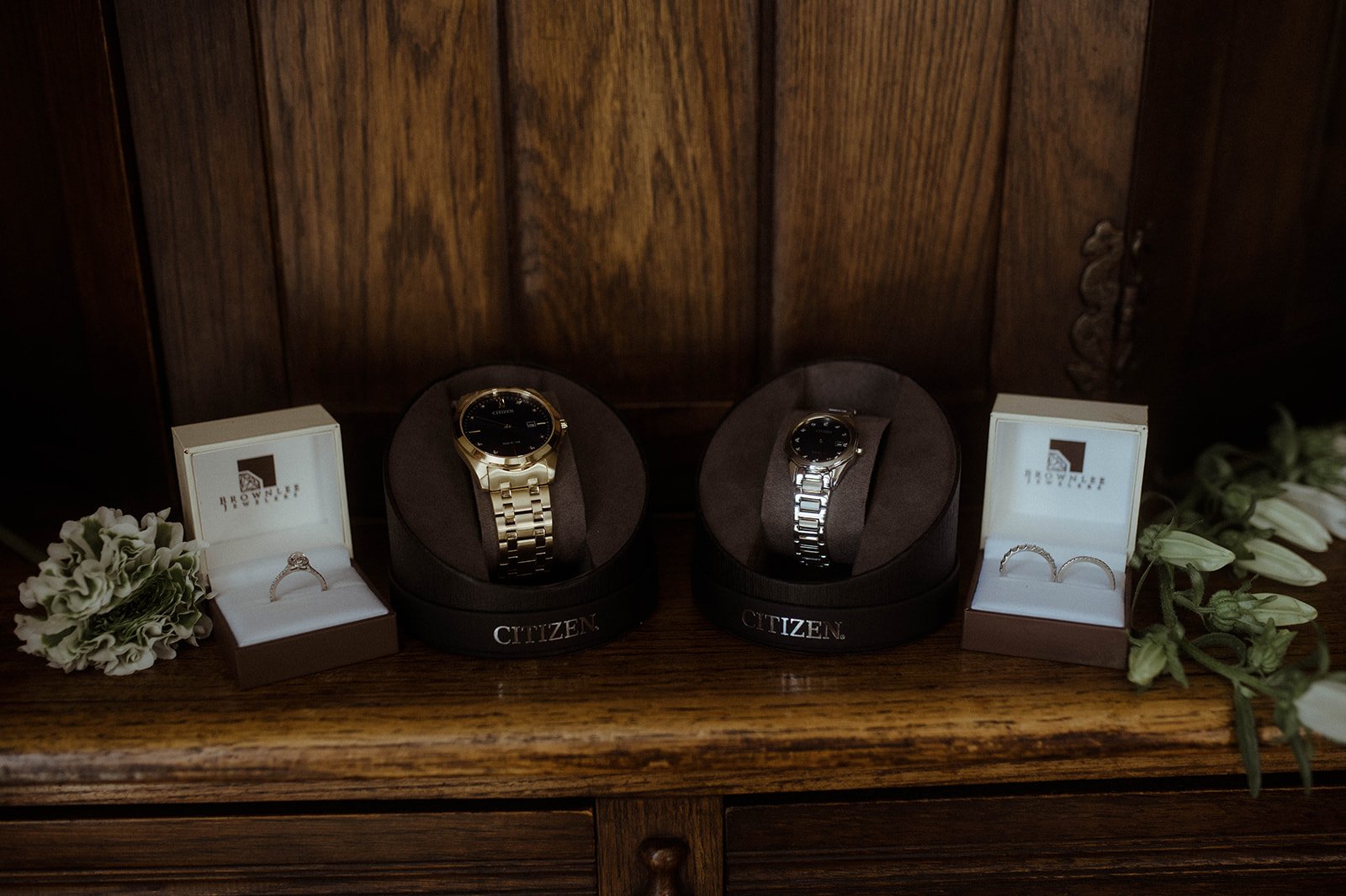 Wedding watches and ring boxes arranged on a wooden cabinet with small greenery accents.