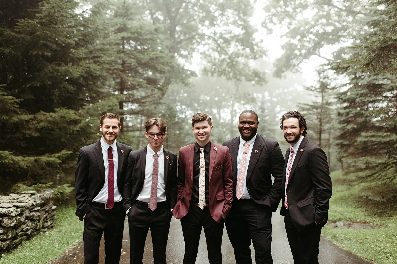 Groom in a burgundy suit posing with groomsmen in black suits during a misty forest wedding portrait.