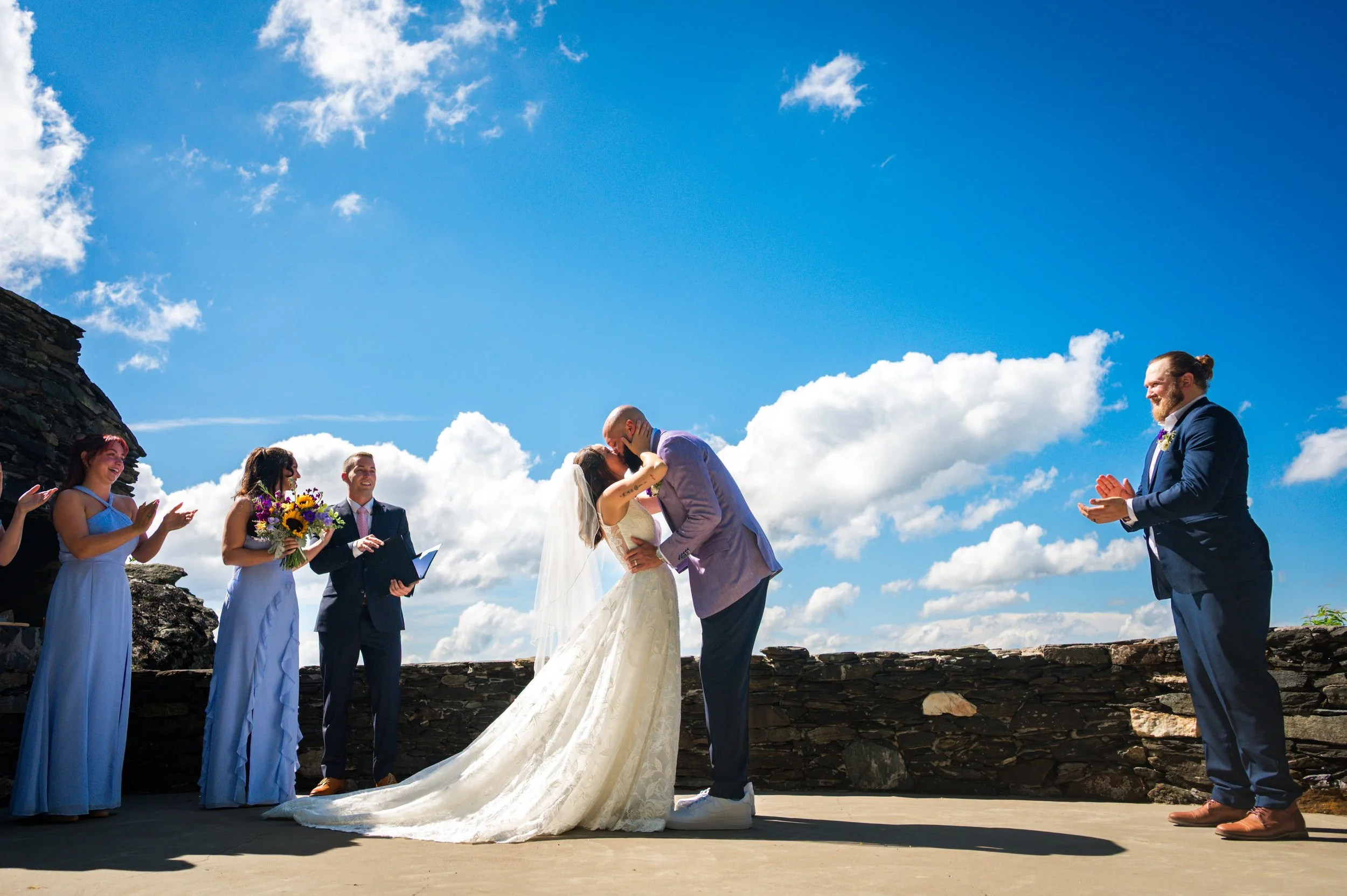 Bride and groom sharing their first kiss during an outdoor mountain wedding ceremony with bridesmaids, officiant, and blue sky backdrop