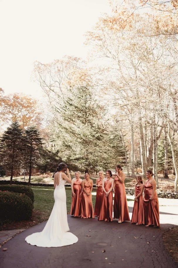 Bride showing her dress to bridesmaids outdoors as they react during a fall wedding