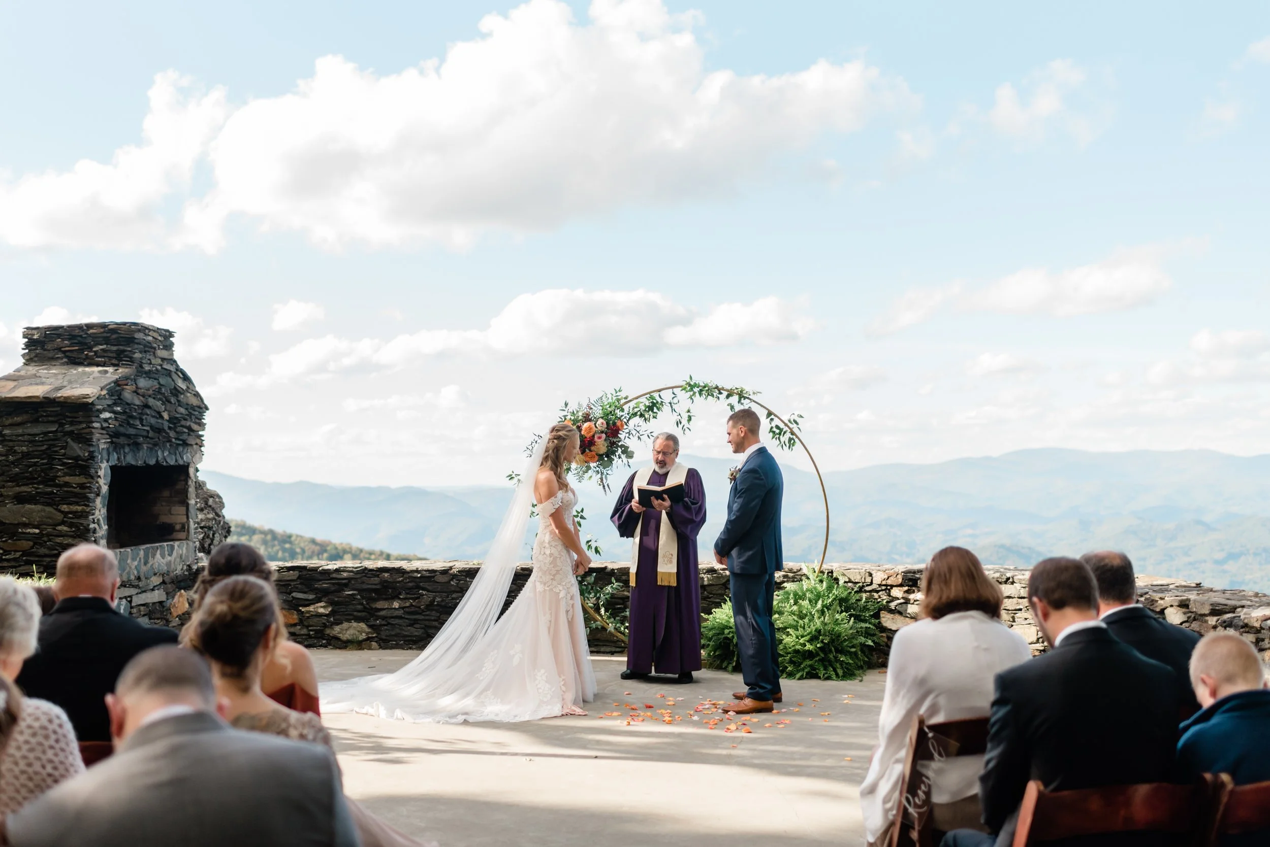 Bride and groom exchanging vows during a mountain overlook wedding ceremony with a circle floral arch and stone fireplace.