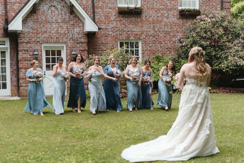 Bride greeting bridesmaids in blue dresses as they walk across a lawn outside a brick estate wedding venue.