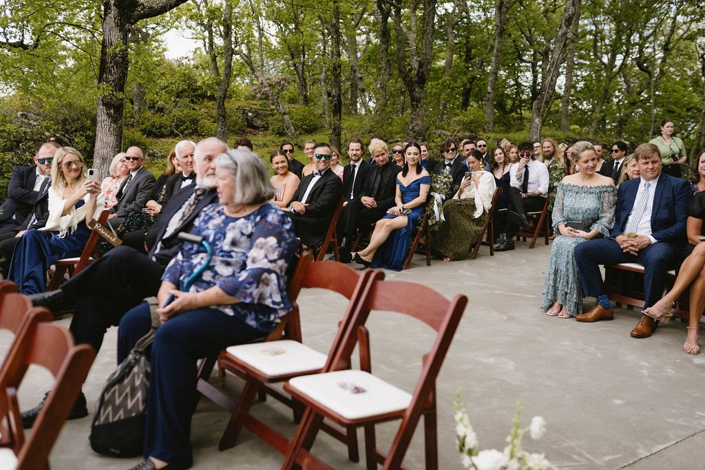 Wedding guests seated in wooden chairs during an outdoor mountain ceremony surrounded by trees and stone walls.